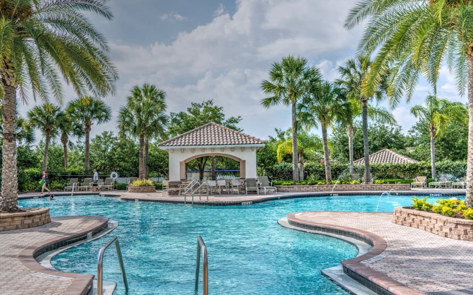 Resort pool deck of interlocking stone pavers with palm planters and a tiled pavilion