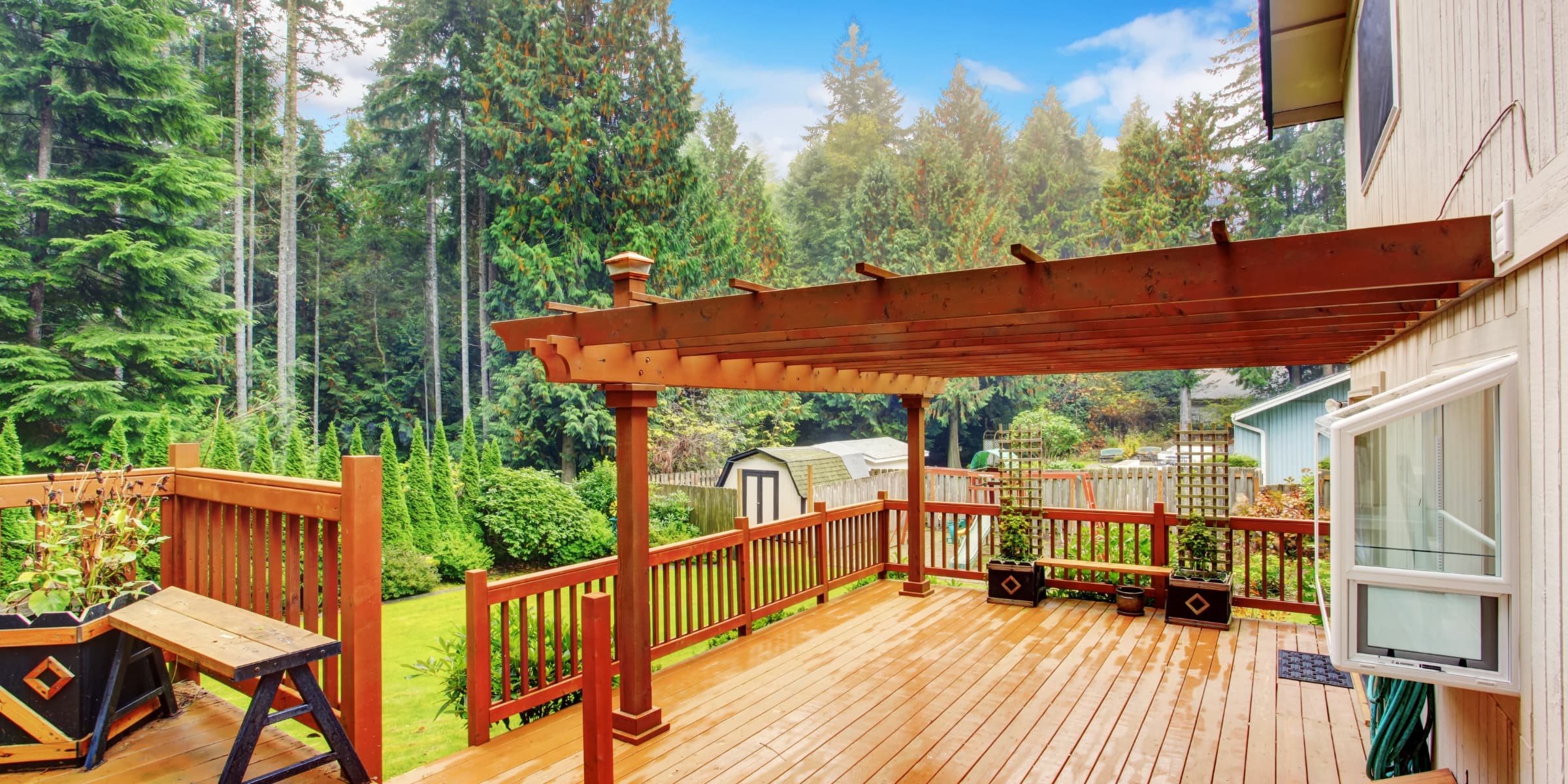 Stained wood deck and pergola beside a house, with wet deck boards and evergreen trees